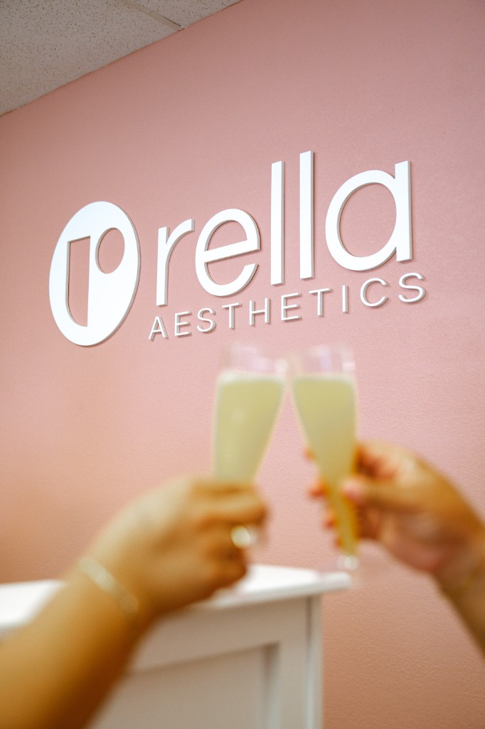 Rella Aesthetics logo on a pink wall, with champagne glasses raised in front